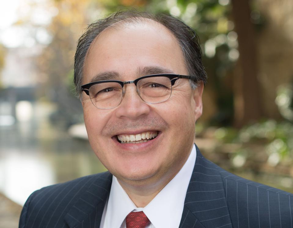Man with glasses smiles, wearing a suit, outdoors with a water and green trees in the background.