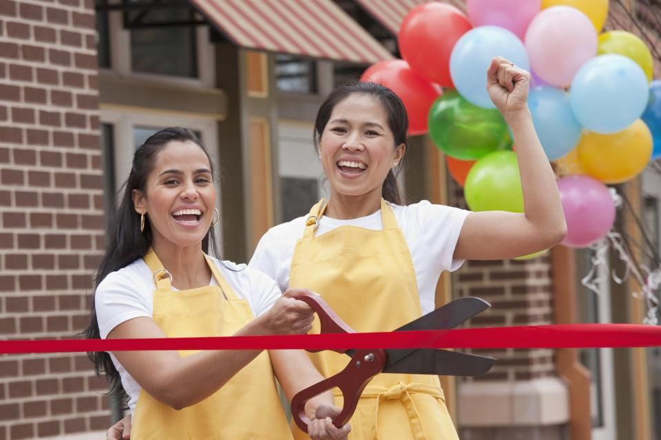 Two women in yellow aprons cutting a red ribbon with scissors in front of a store with balloons.