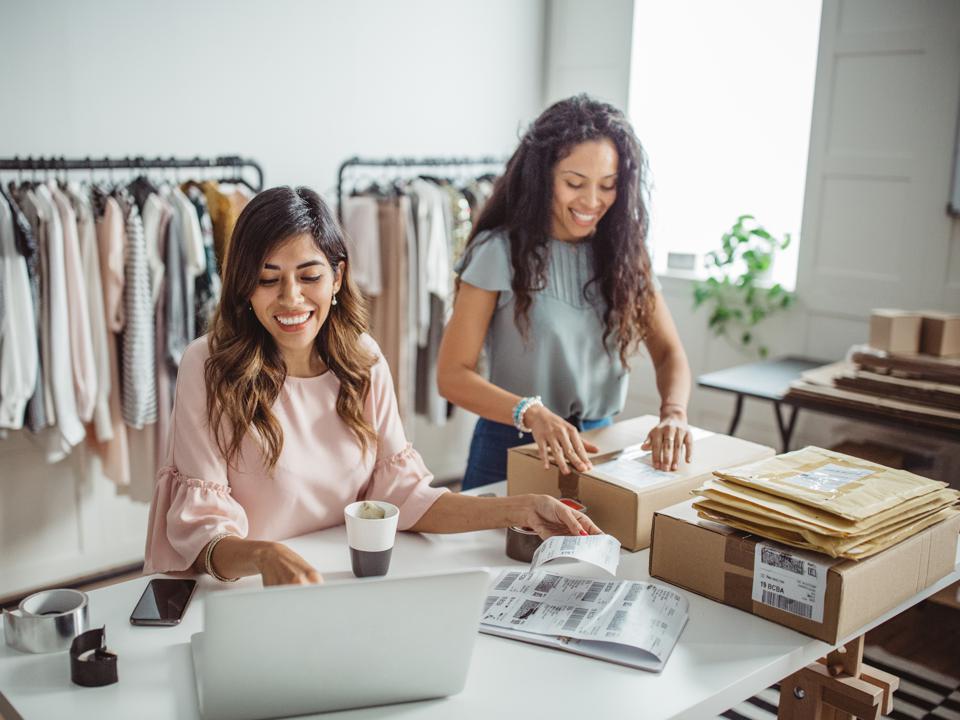 Two women packing boxes at a desk, surrounded by clothes racks, smiling and working.