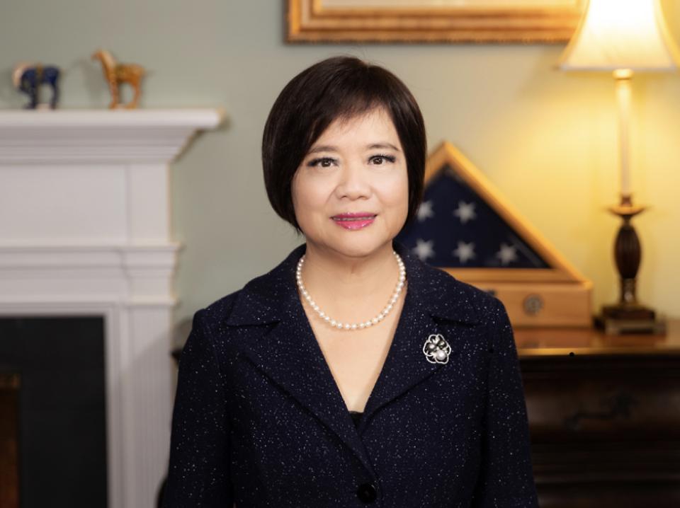 Woman in navy suit with a pearl necklace, smiling, indoors. A flag and lamp are visible in the background.