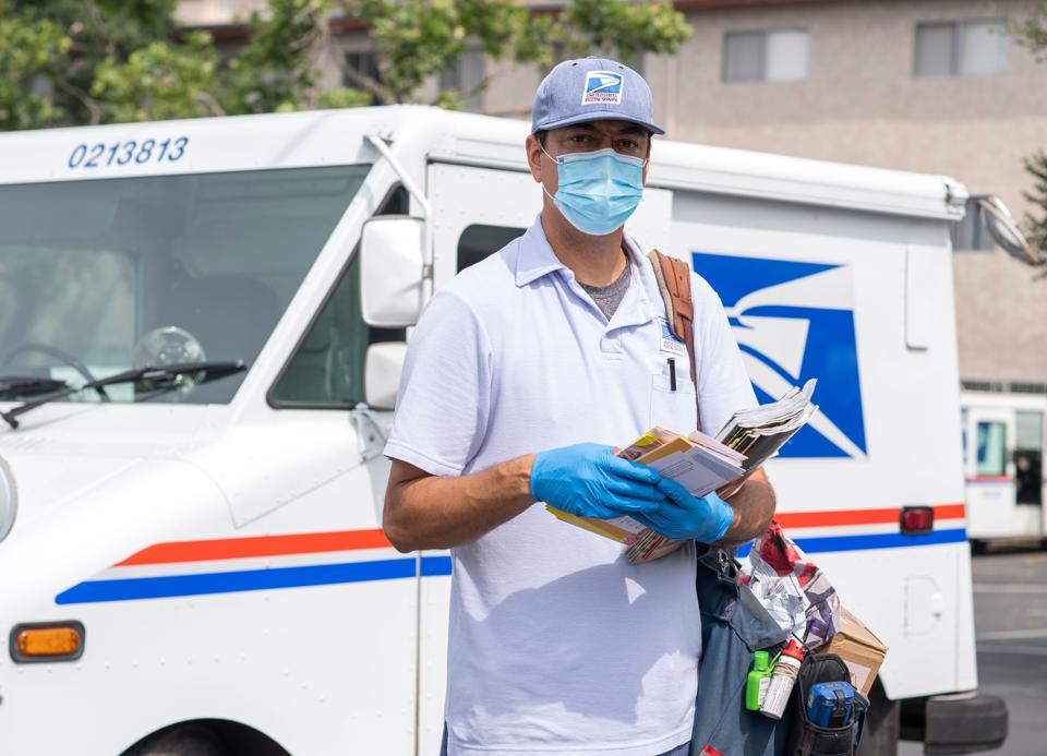Postal worker wearing a mask and gloves, standing near mail truck, holding mail.