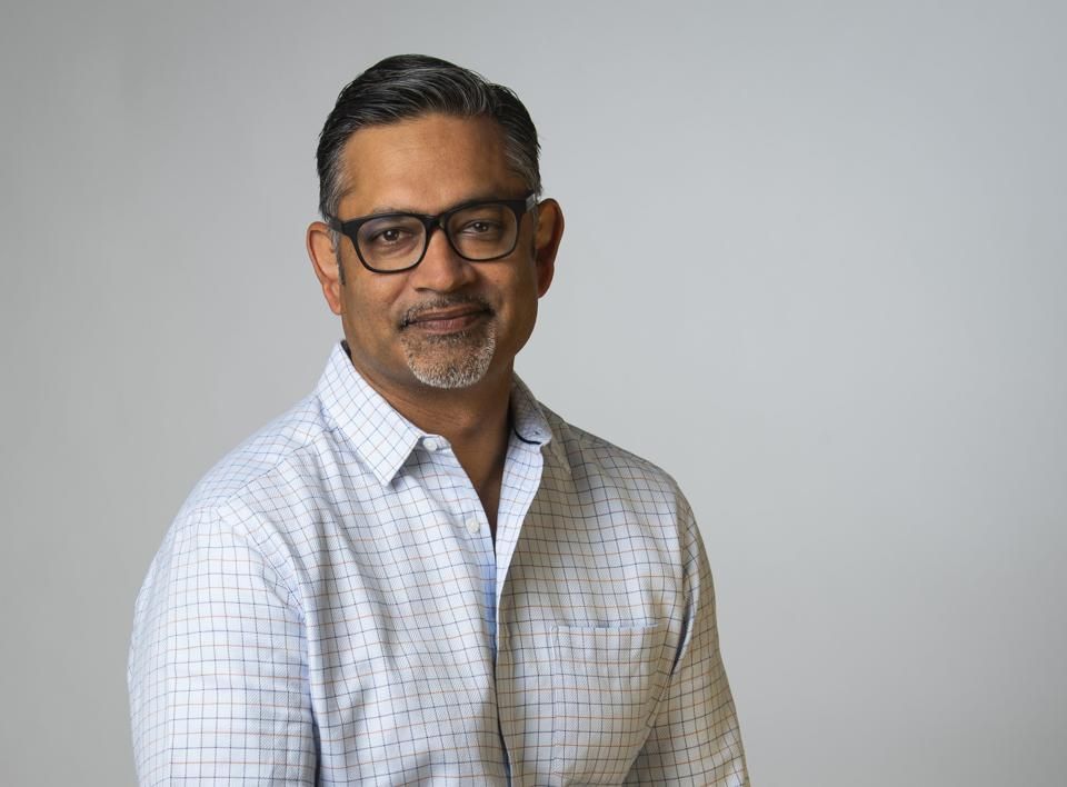 Man with glasses, wearing a light blue checked shirt, smiling, against a gray backdrop.