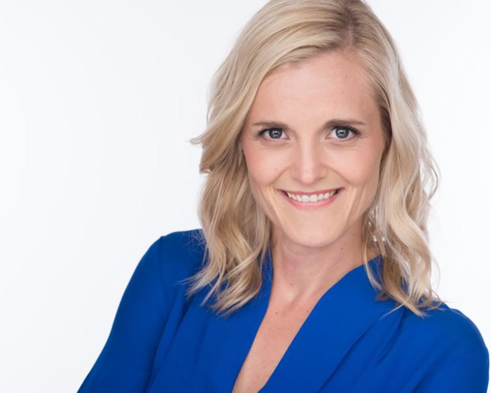 Blond woman in blue top smiles at camera, studio shot with white background.