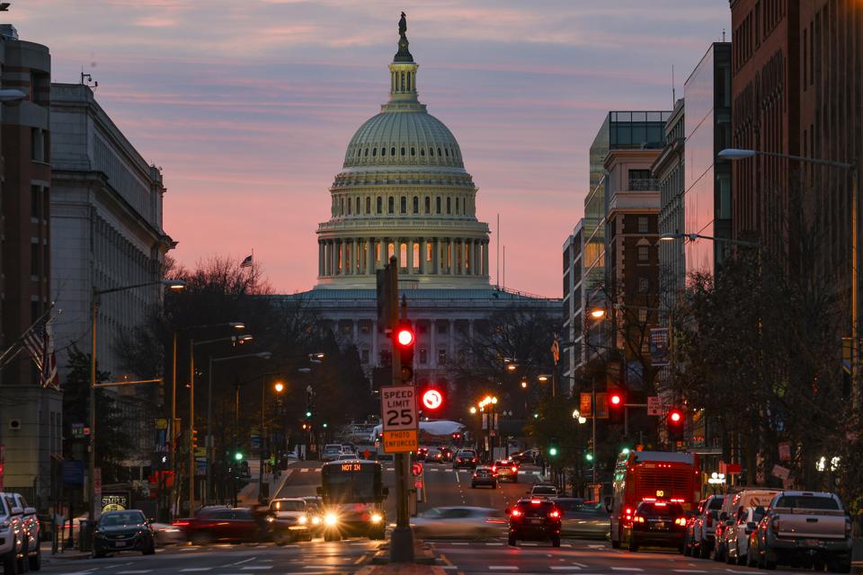 U.S. Capitol building at dusk, viewed down a city street lined with cars and buildings under a pink and orange sky.