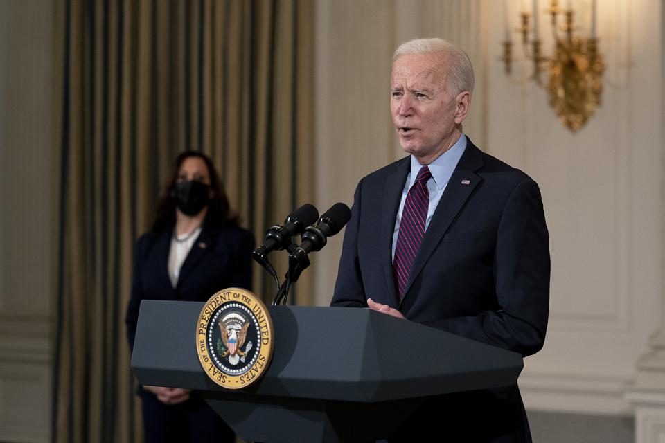 President Biden speaking at a podium with Vice President Harris wearing a mask in the background.