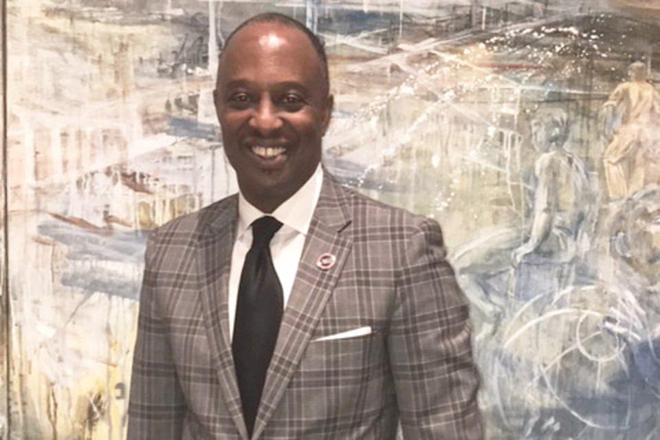 Man in gray plaid suit, smiling, in front of abstract artwork.