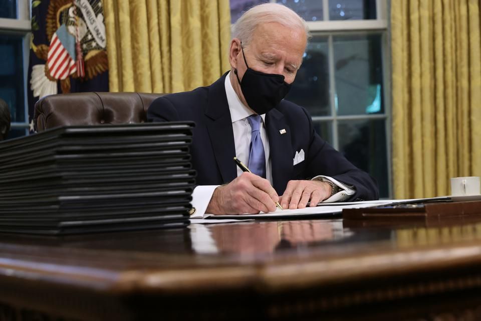 President Biden in the Oval Office, signing documents at a desk, wearing a mask.