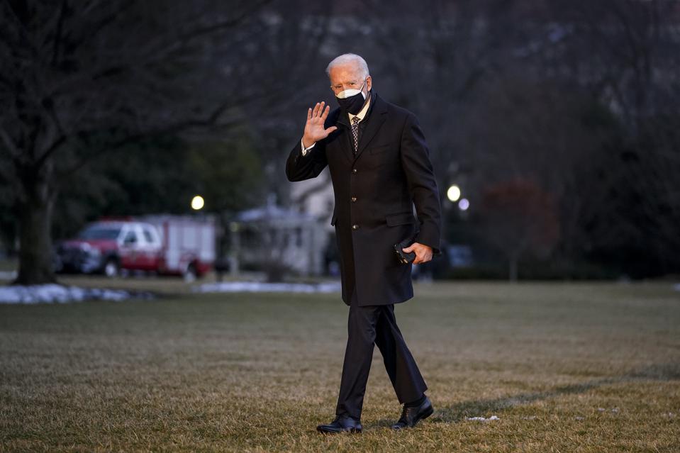 Joe Biden in a black coat and mask waves on a grassy lawn.