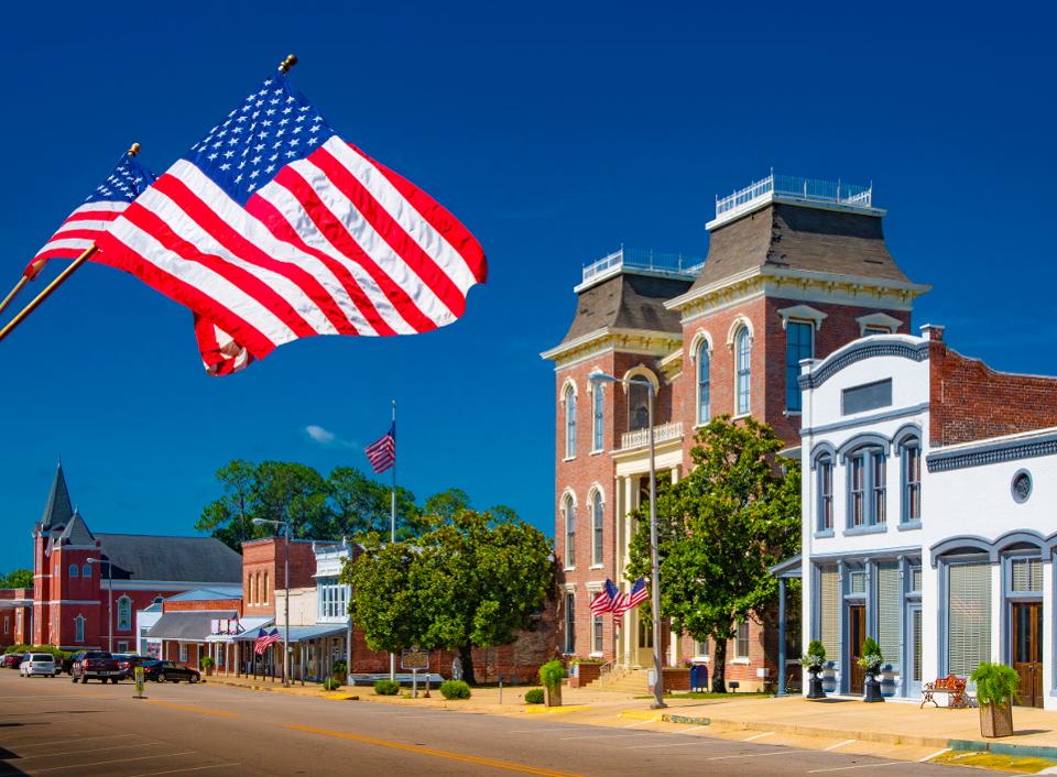 American flags fly over a historic downtown street with brick buildings on a bright, sunny day.