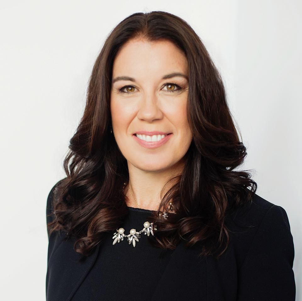 Woman with dark wavy hair smiles, wearing a black blazer and necklace, against a white background.