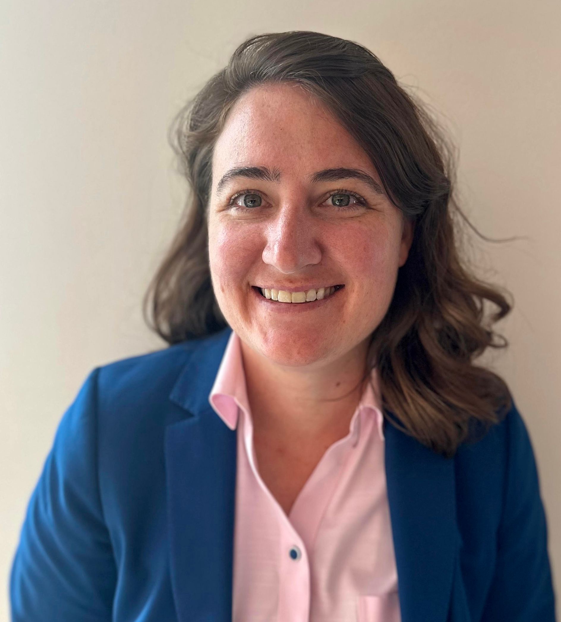 Woman with brown hair smiles, wearing a pink shirt and blue blazer against a light wall.