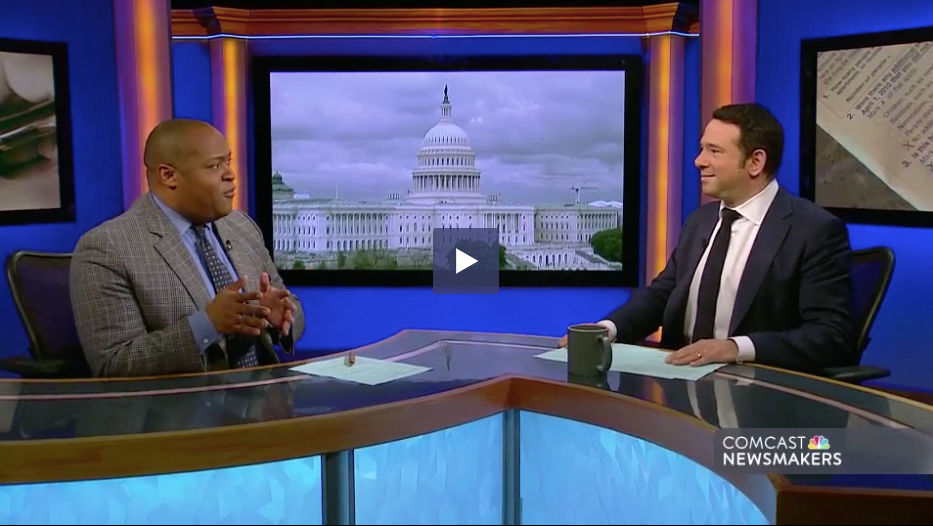 Two men in suits at a news desk. US Capitol background. One man gestures with hands while speaking, other smiles.