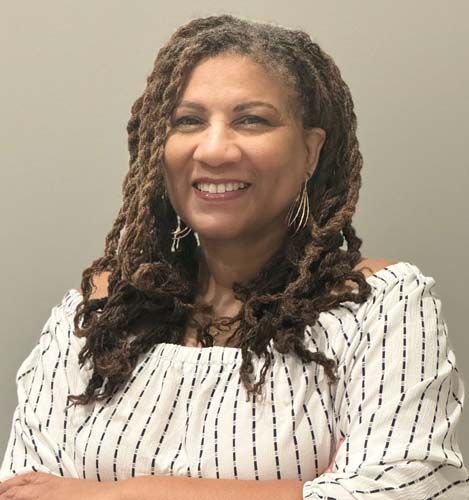 Woman with long, brown locs smiles, wearing off-shoulder white top with vertical black stripes; against a gray wall.