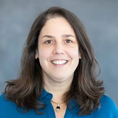 A woman wearing a blue shirt and a necklace is smiling for the camera.