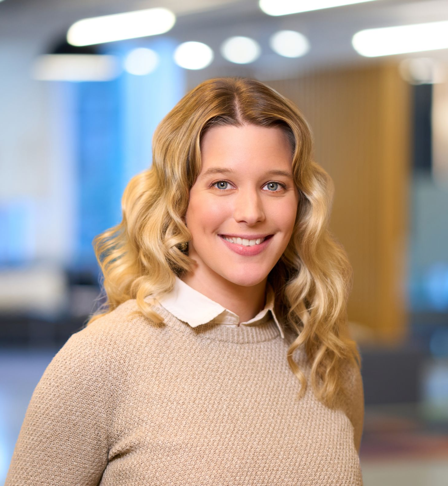 Blonde woman smiling, wearing a tan top, against a blurred office background.