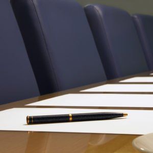 Pen on a white table in front of a row of blue chairs, in a bright conference room.