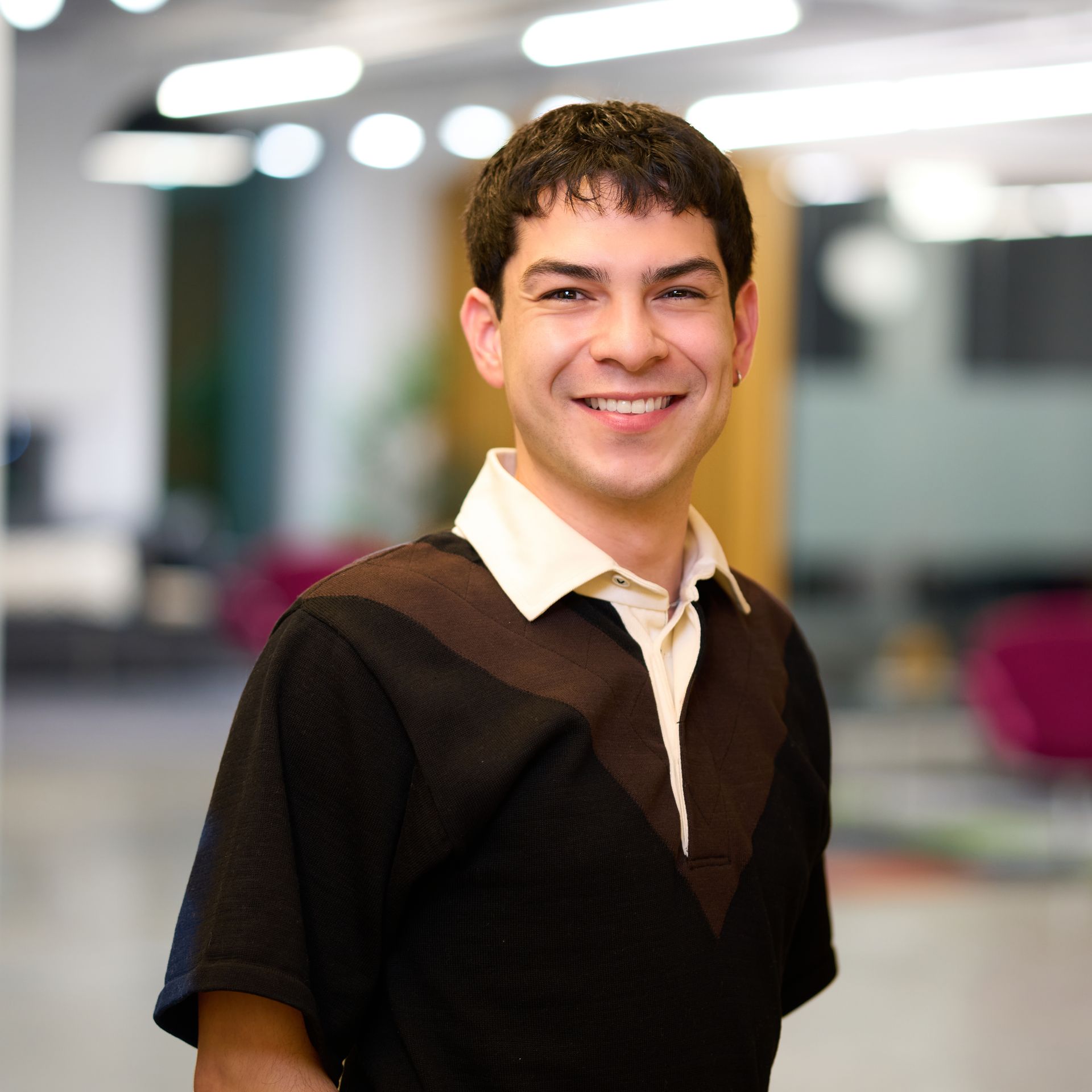 Man smiling, wearing a black and brown shirt against an office background.