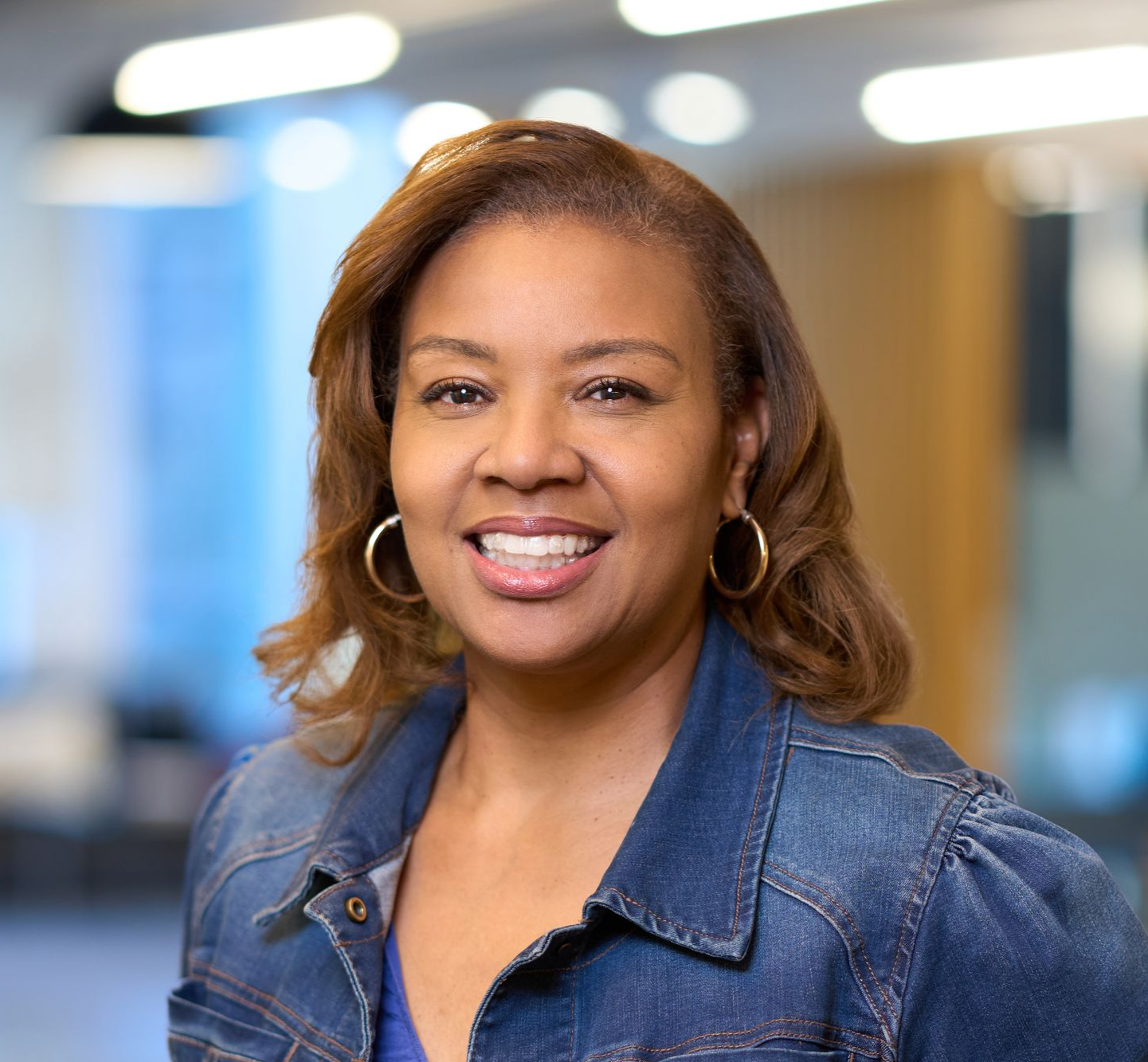 Woman in denim jacket smiles in front of blurred office background.