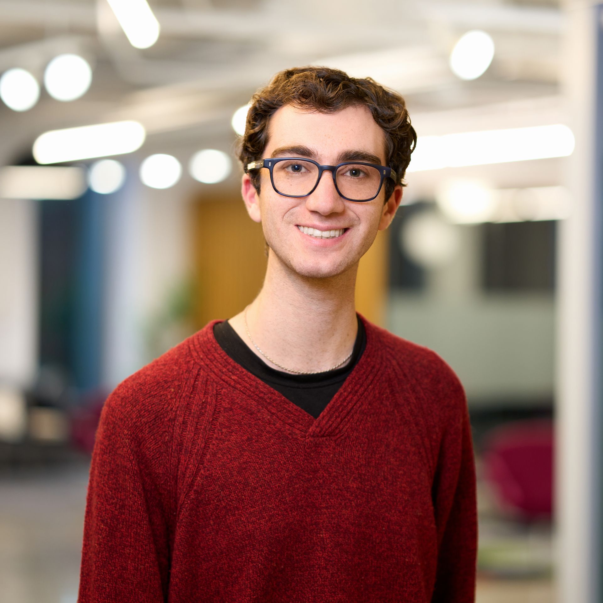 Man in red v-neck sweater smiles in front of a blurred office background. 