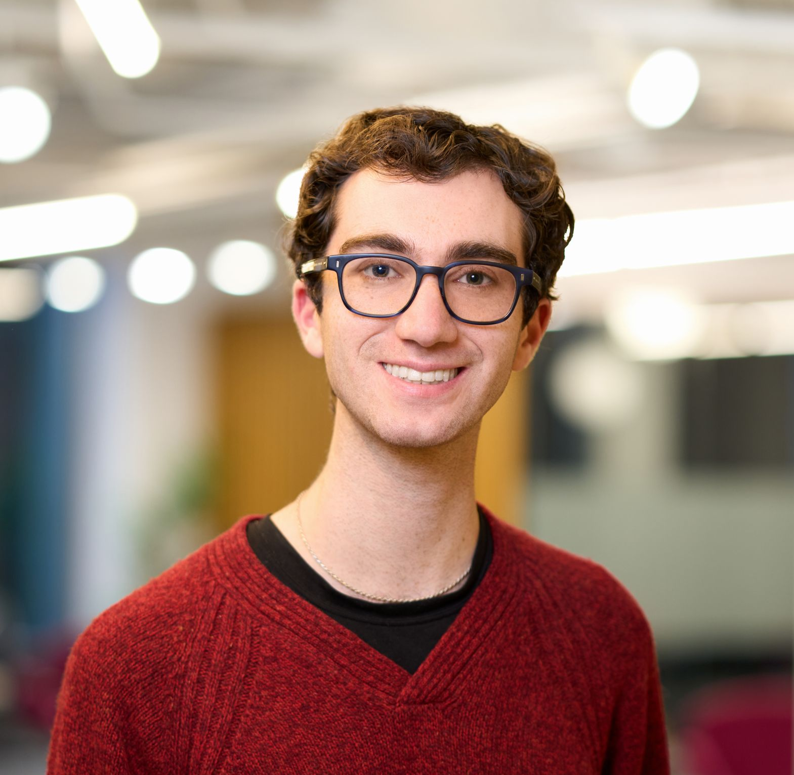 Man in glasses smiling, wearing a red v neck sweater against an office background.