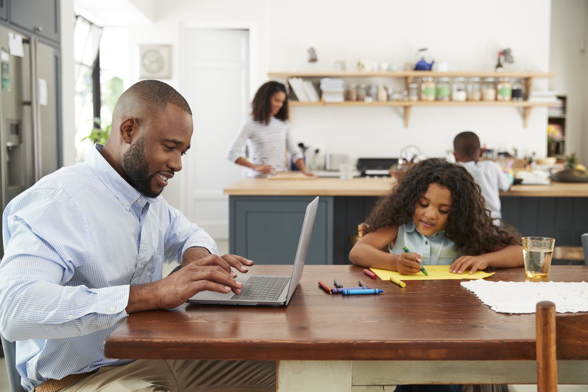 A man is sitting at a table using a laptop while a little girl does her homework.