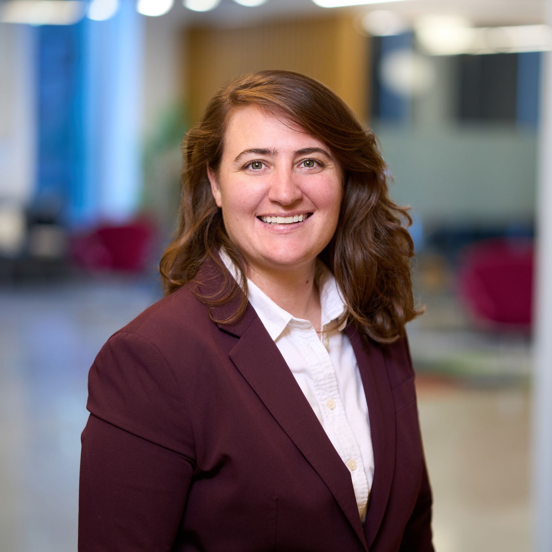 Woman with brown hair smiling, wearing a blue blazer over a pink collared shirt. Against a neutral wall.