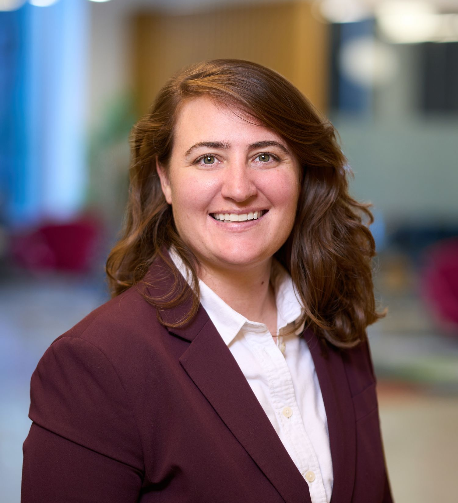 Woman with brown hair smiles, wearing a white shirt and burgundy blazer against a blurred background.