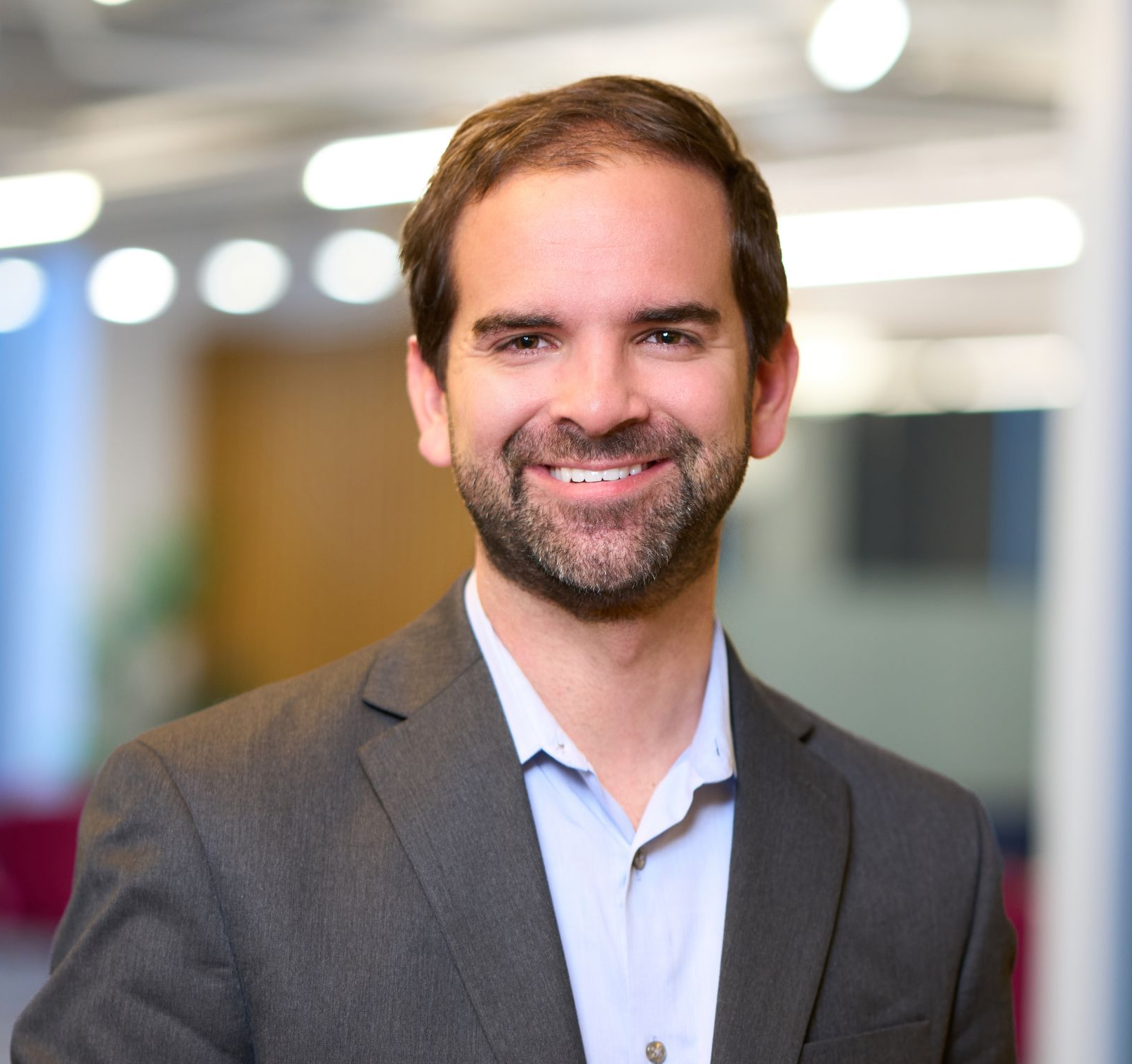 Man with a beard in a blue shirt and dark blazer smiles, against a blurred office background.