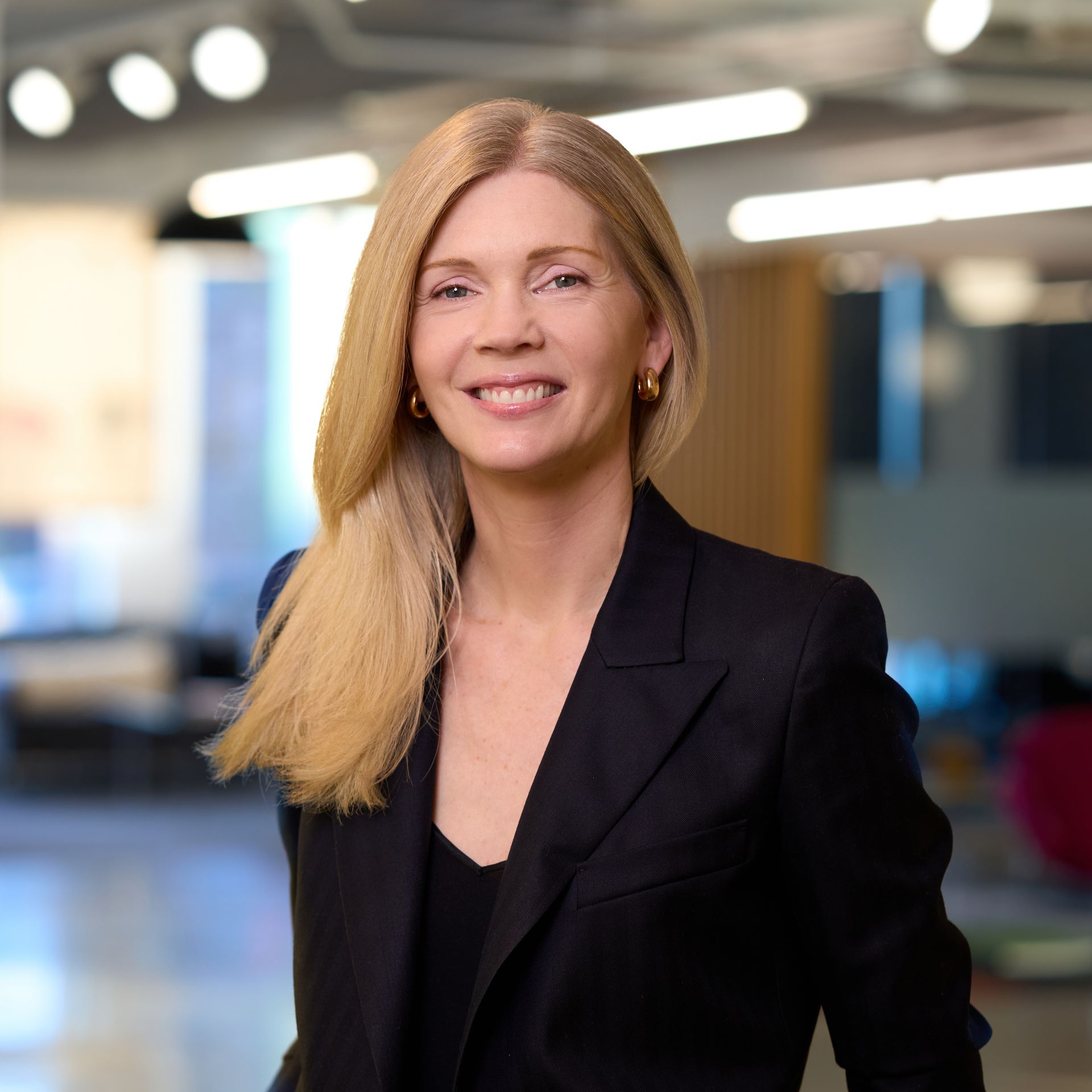 Blond woman in dark blazer smiles, soft background.