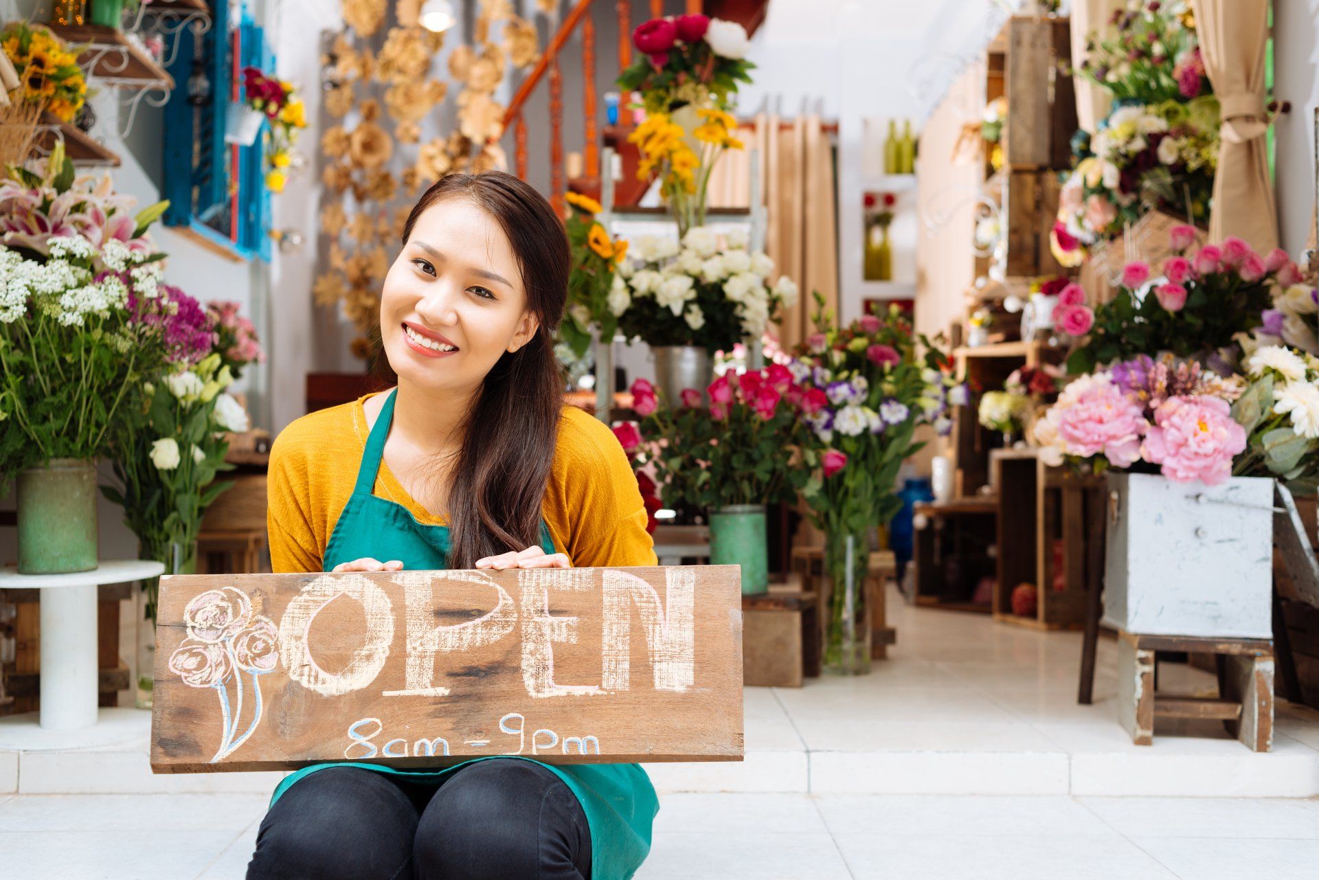 A woman is sitting in front of a flower shop holding an open sign.