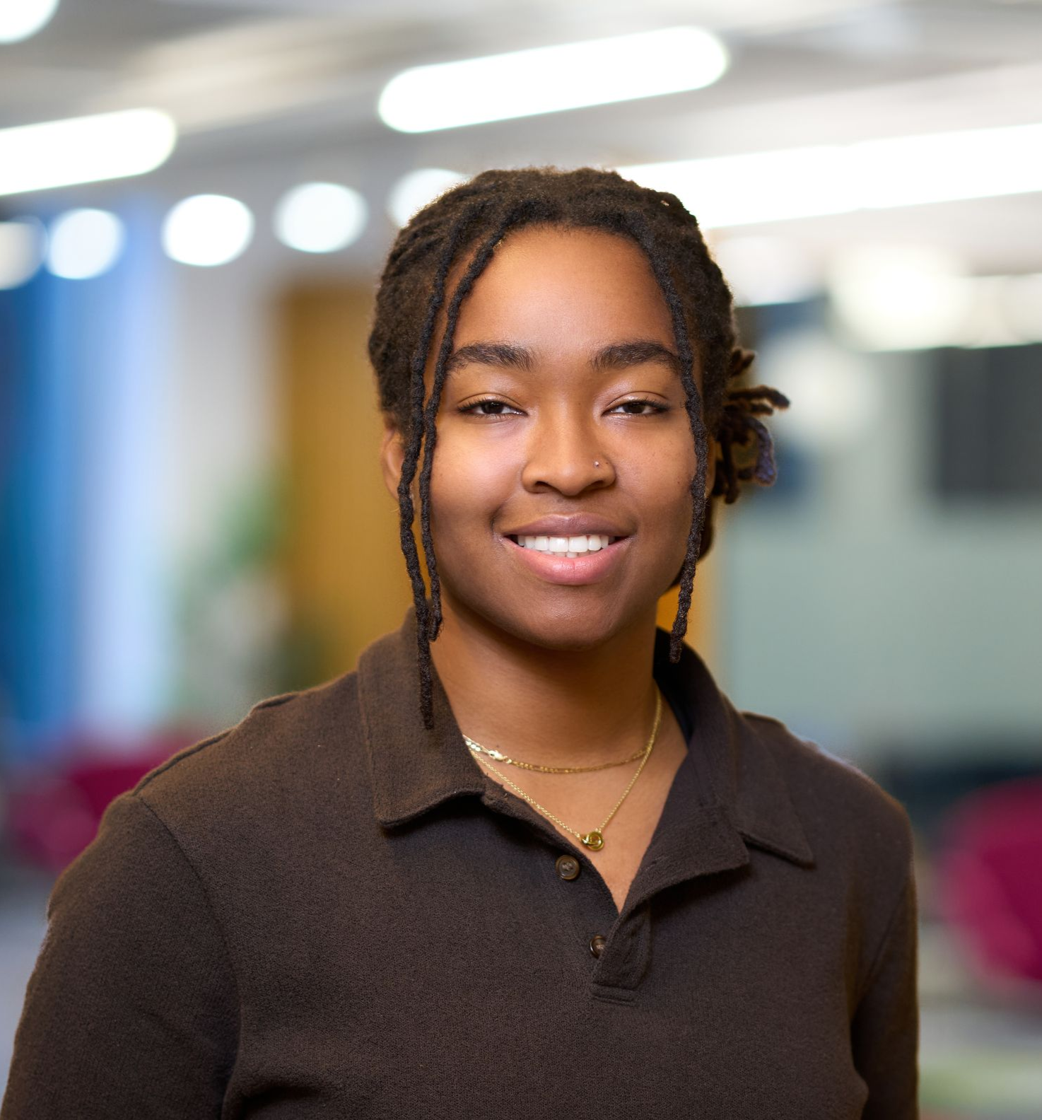 Woman with locs smiling, wearing a brown shirt, against a blurred office background.