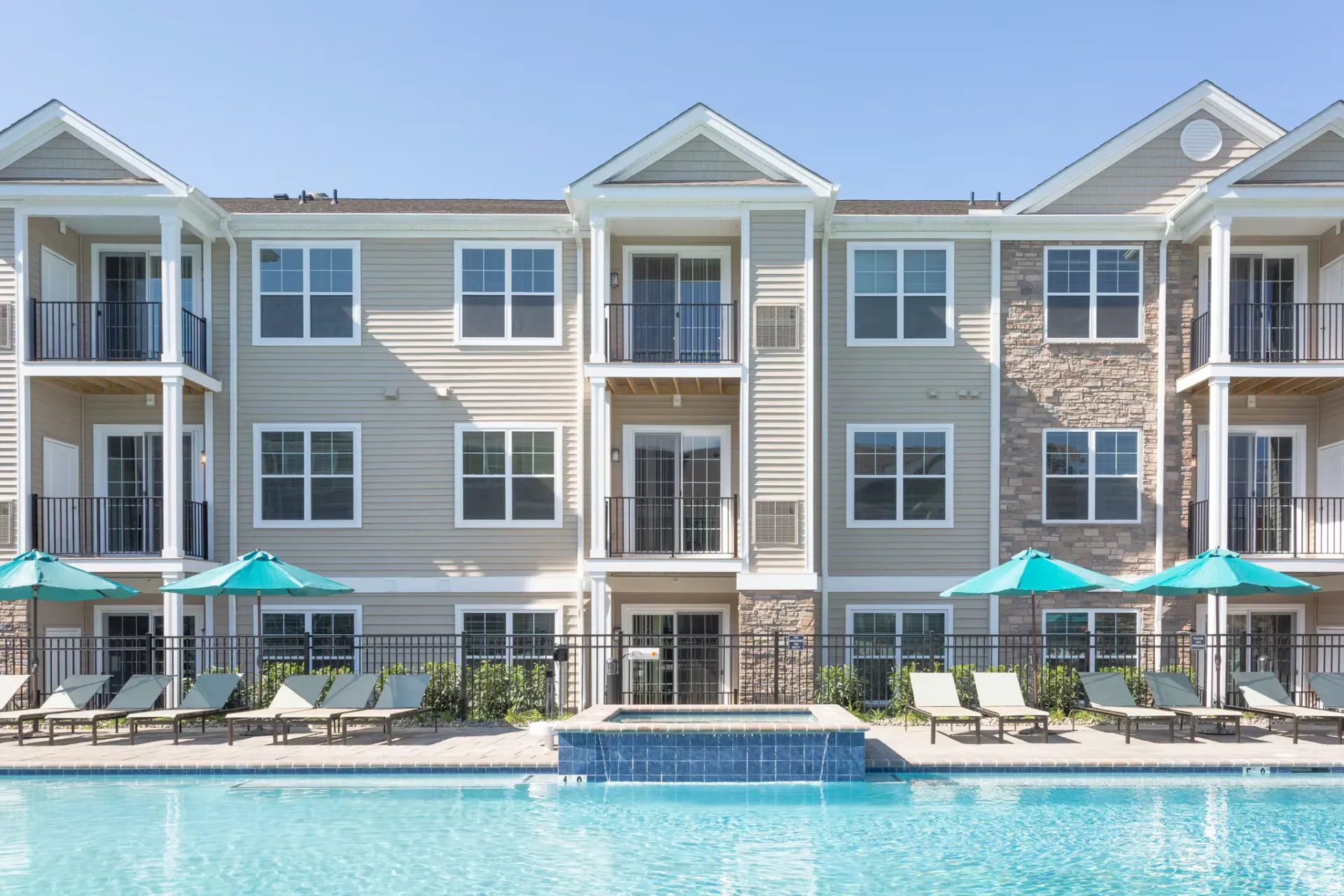 Apartment building exterior with balconies, pool, and lounge chairs on a sunny day.