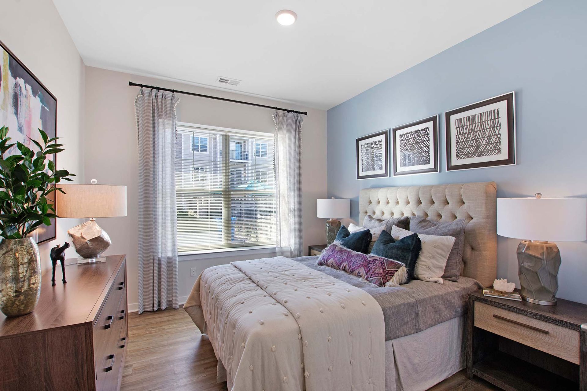 Bedroom with tufted beige headboard, layered bedding, nightstands, and a window with sheer curtains at The Parc at Roxbury in  Roxbury, NJ.