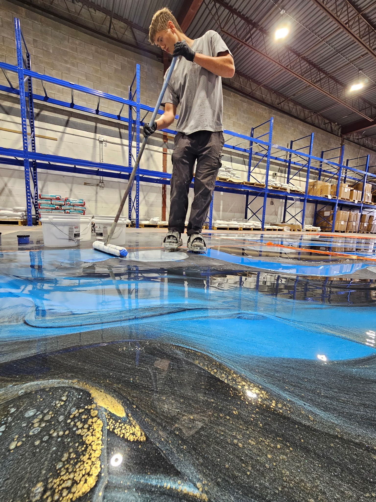 Personne en chemise et pantalon gris appliquant de l'époxy bleu sur le sol d'un entrepôt.