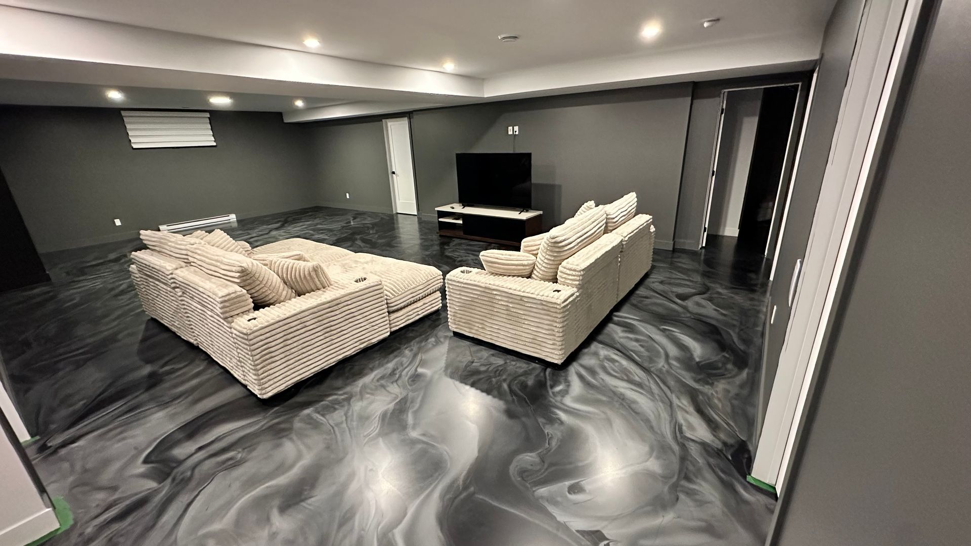 Salon moderne au sous-sol avec deux canapés beiges, télévision et sol en marbre foncé