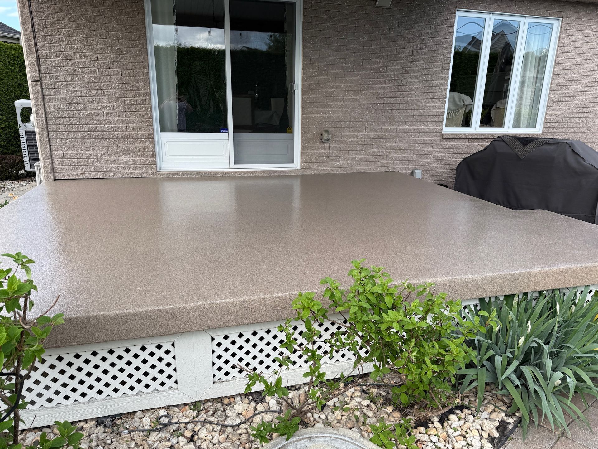 Patio en béton beige, maison en brique, porte coulissante en verre, fenêtre, grille couverte, arbustes au premier plan.