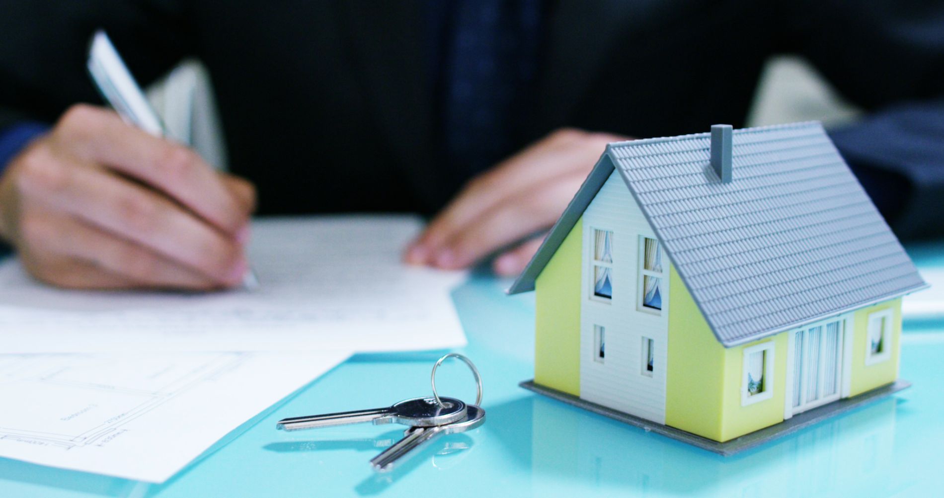 a blurred image of someone signing a property contract with a miniature house and a set of keys on a desk in the foreground