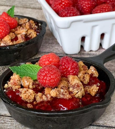 granola crumble in mini cast iron skilled topped with raspberries by a bowl of fresh raspberries in white container