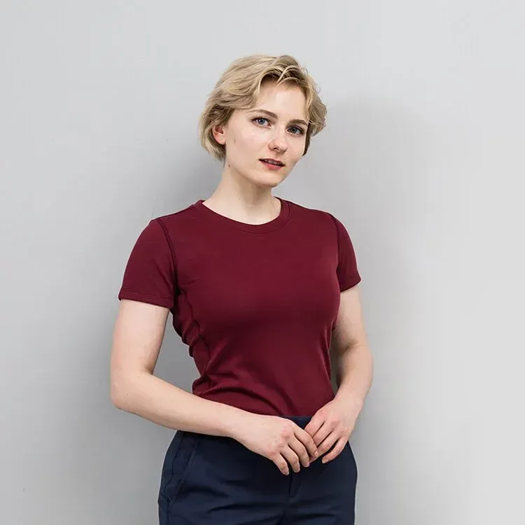 一位留著金色短髮的女子，身穿栗色襯衫和藏藍色褲子，站在一面灰色牆前。