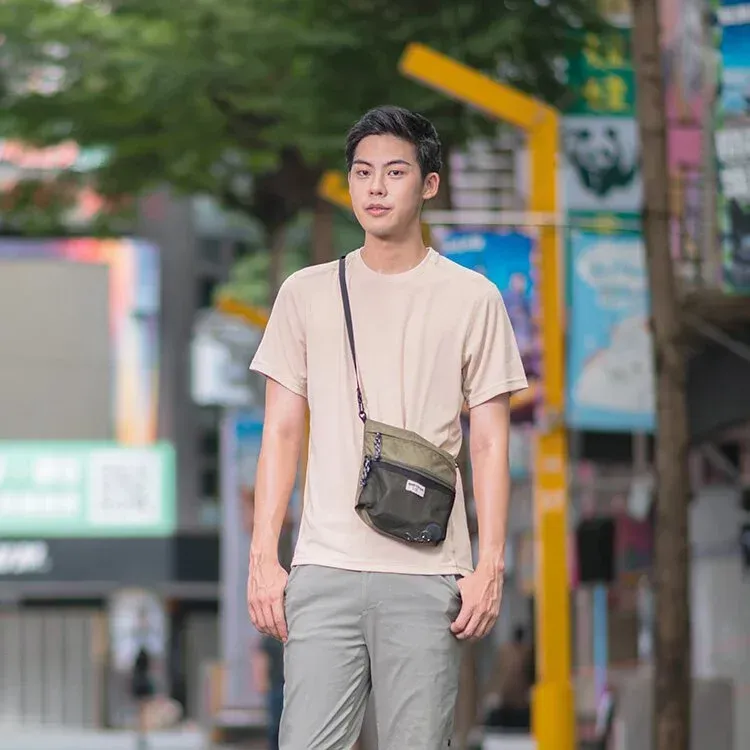 一名身穿灰色 T 恤和黑色褲子的男子站在那裡，雙手插在口袋裡。