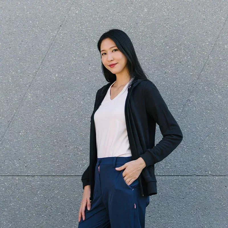一位身穿黑色夾克和藍色褲子的女子站在一堵灰色的牆前。