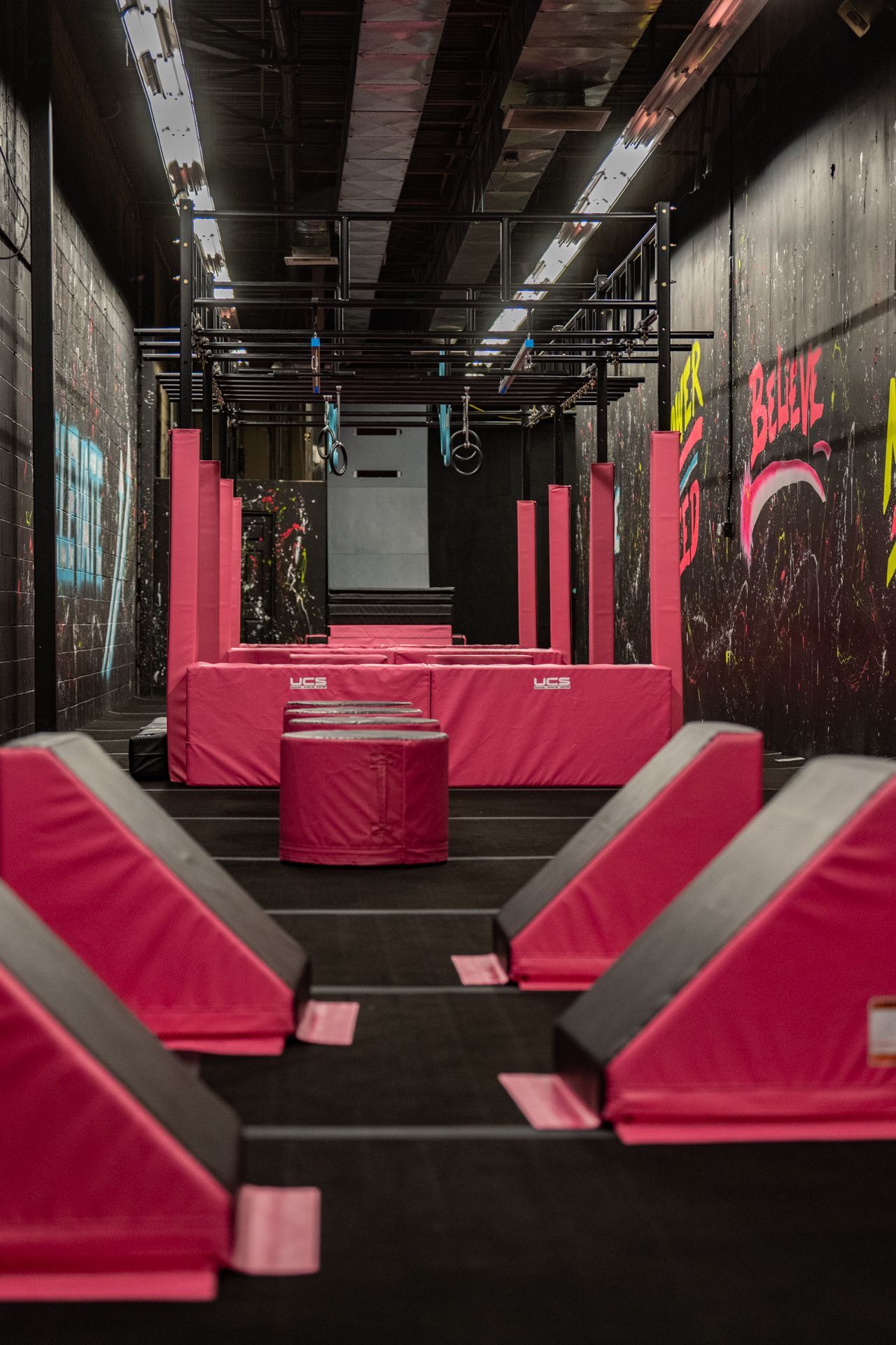 Indoor parkour gym with pink obstacles and overhead bars, dark walls, and black flooring.