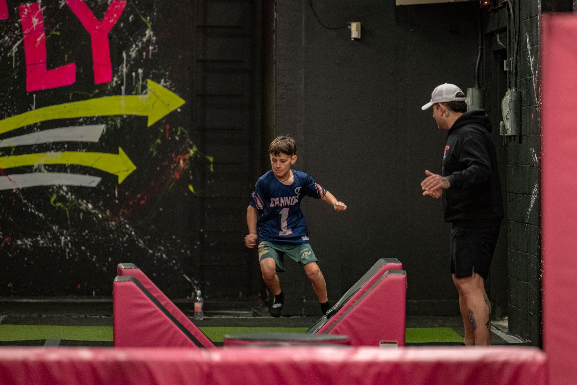 Boy jumping over obstacles in an indoor parkour gym, with an instructor watching.