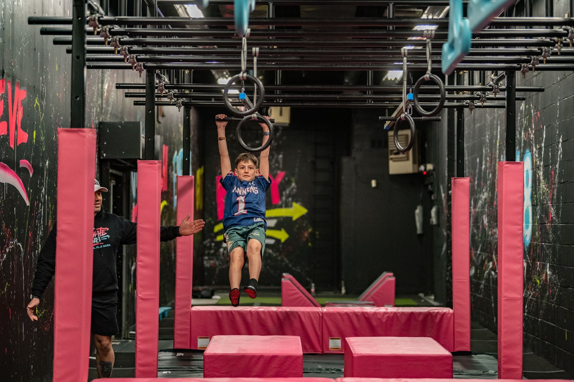 Boy hanging from rings on an obstacle course, instructor watches. Pink and black setting with graffiti.