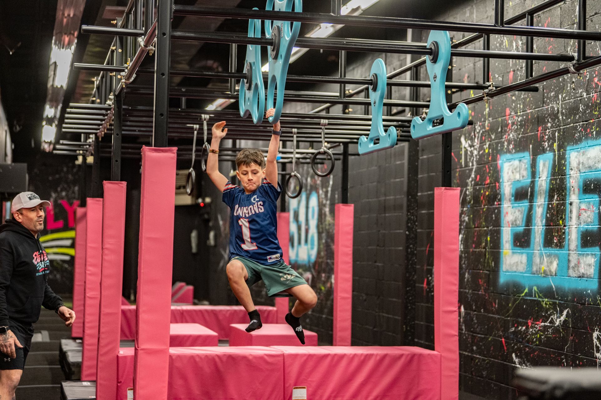 Boy swinging on obstacle course bars, pink mats, black walls, instructor watching.