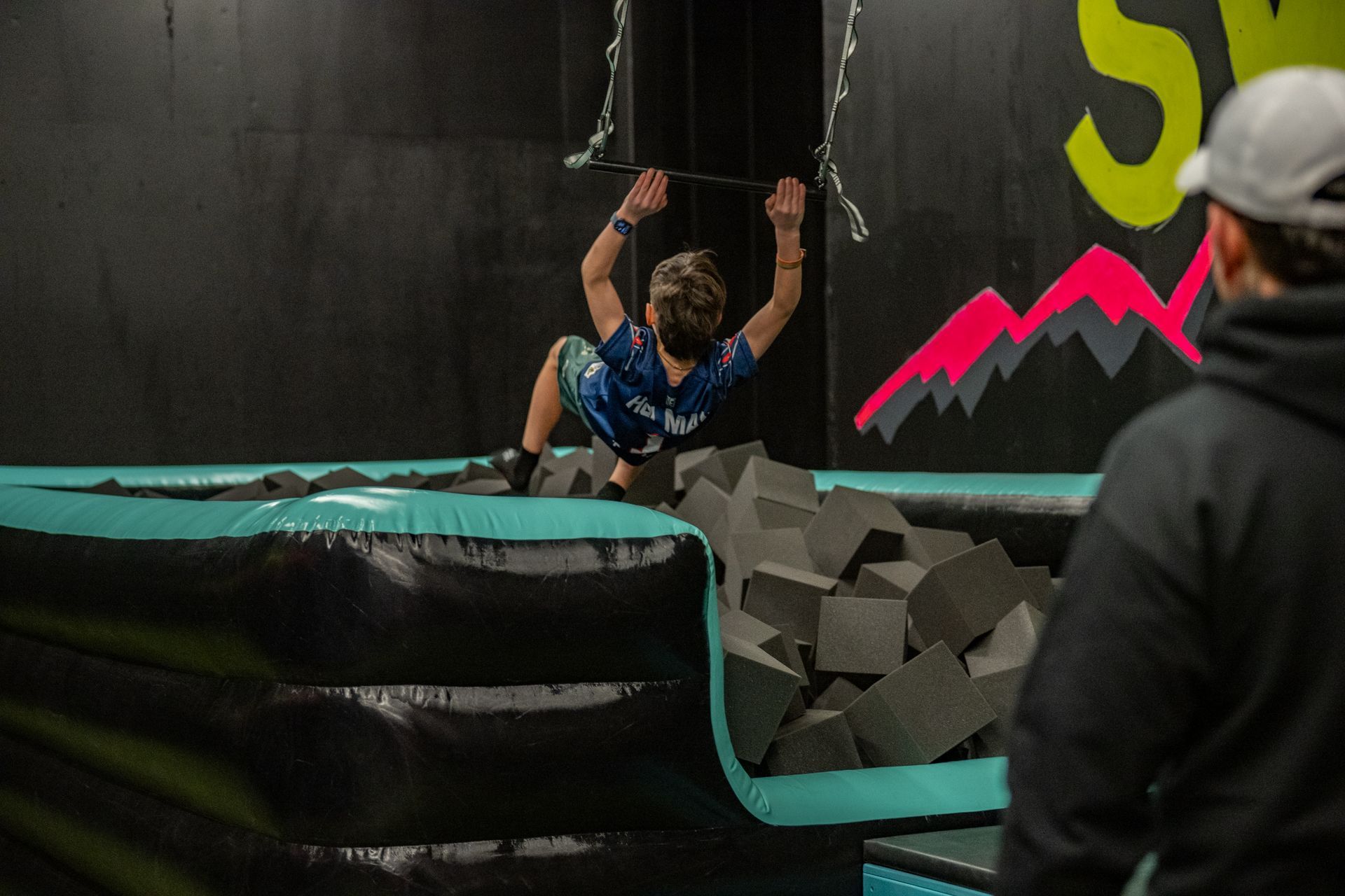 Boy swinging on rope over foam pit in an indoor trampoline park; instructor watches.