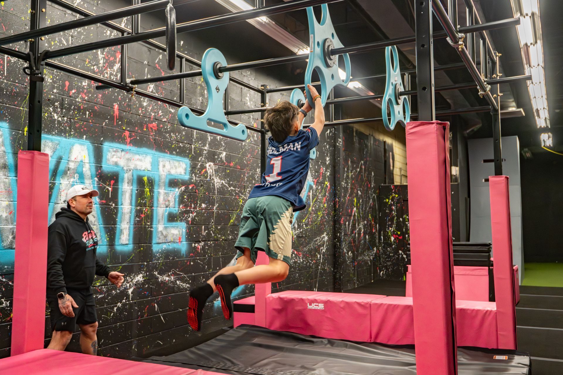 Boy on an obstacle course, hanging from grips. A coach watches. Indoor gym with pink and black elements.