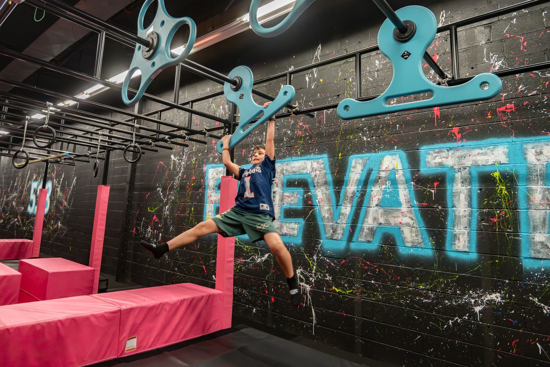 A person swinging on a ninja course, reaching for a blue handle. Black and pink walls.