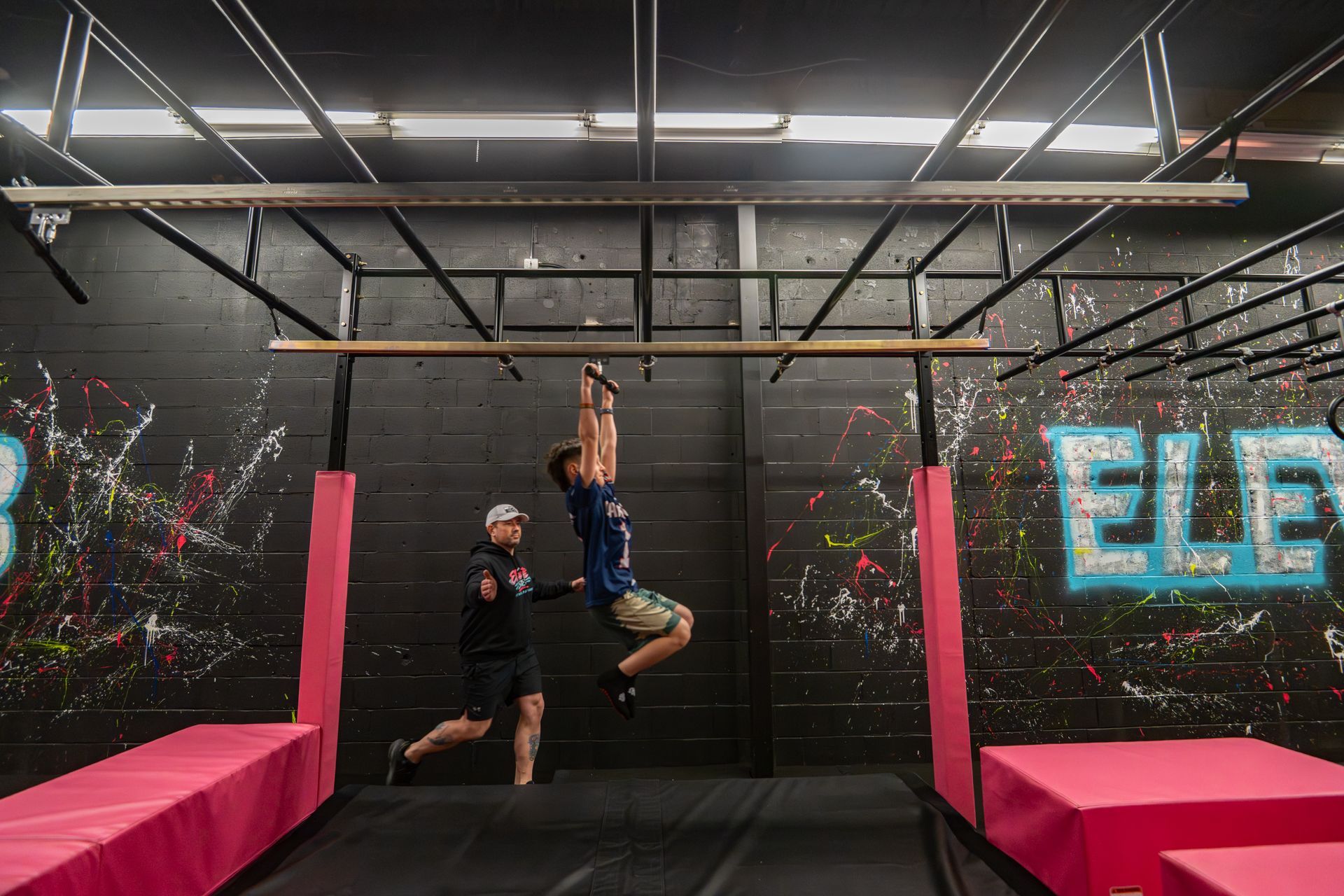 Man swinging on a bar, aided by a person on the ground in a gym with pink mats and a colorful wall.