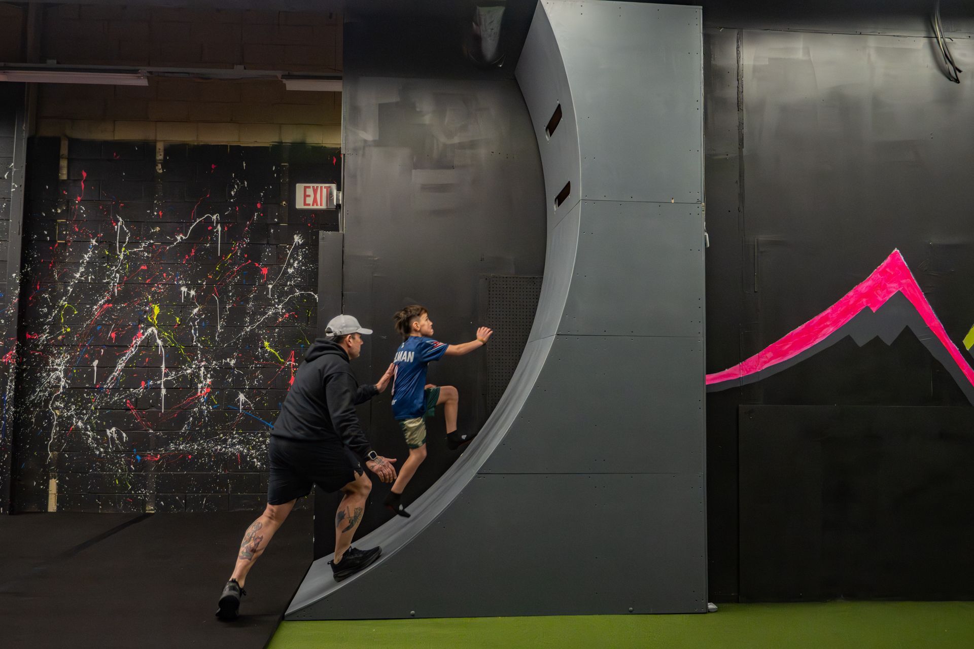 A person coaches a child climbing a curved wall at an indoor obstacle course.